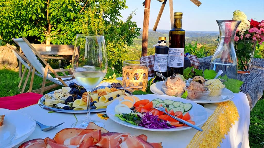 Das Bild zeigt einen gedeckten Tisch auf der Sonnenterrasse mit hausgemachten Köstlichkeiten wie Schinkenteller, Gemüseteller, Essig und Kernöl, Aufstriche sowie einen Käseteller. Im Hintergrund erkennt man die südsteirischen Weinberge.